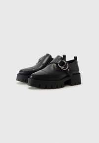 Black leather shoes with a chunky sole, featuring a wide strap and circular silver hardware accent. Smooth texture with stitching details.