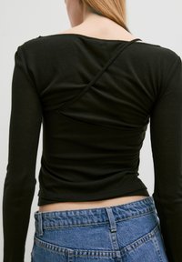 Schwarzes geripptes Langarmoberteil mit einem runden Rückenausschnitt und überkreuzten Trägern. Kombiniert mit hochgeschnittenen blauen Jeans.