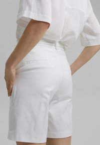Personne portant un short blanc taille haute et une chemise blanche à manches courtes ample, debout avec les mains posées sur les hanches.