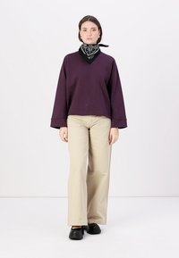Felpa viola over size con scollo a V, abbinata a pantaloni beige a gamba dritta. Bandana nera attorno al collo e calzature nere.