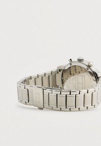 Ezüst fém láncóraszíj csattal, rajta gravírozott logó, a kerek karórához rögzítve.