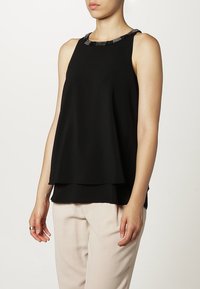 Svart ärmlös topp med ett lagerdesign och en dekorativ pärlkrage. Mjuk, slät tyg med lös passform, ihop med ljusa byxor.