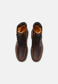 Brown leather lace-up boots with black accents, orange inner lining, contrasting stitching, and a rugged design suitable for outdoor wear.