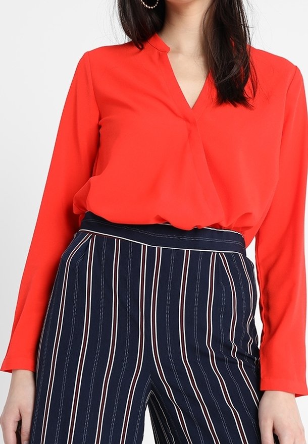 Blouse rouge à manches longues et col en V, associée à un pantalon bleu marine à rayures avec des lignes blanches verticales. Tissu lisse, coupe moderne.