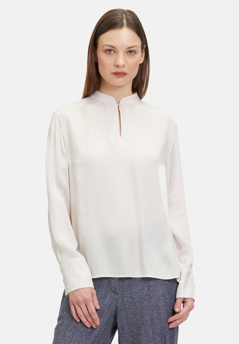 Cremfarbene Bluse mit hohem Kragen, Schlüsselloch-Ausschnitt und langen Ärmeln. Glatte Textur mit dezenter glänzender Oberfläche. Kombiniert mit gemusterten Hosen.