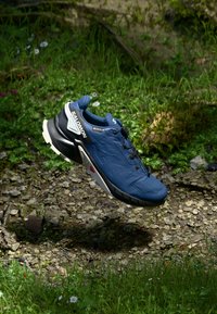 Chaussure de course bleue respirante avec une tige texturée, des accents noirs, un logo blanc proéminent, des détails réfléchissants et une semelle rembourrée, flottant au-dessus de l'herbe.