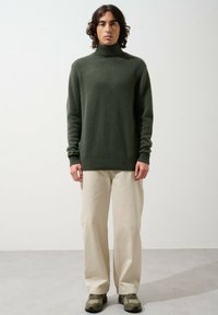 Dunkelgrüner Rollkragenpullover mit langen Ärmeln, kombiniert mit beigen Hosen und grünen Sneakern. Glatte Textur, lockere Passform.
