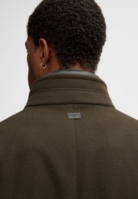 Olijfgroen, wollen mix jas met een hoge kraag, met een label aan de achterkant en een gladde textuur. Het ontwerp is minimalistisch en modern.