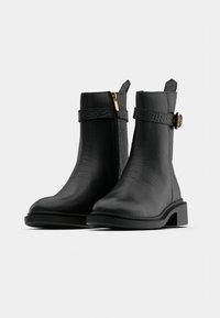 Pár černých kotníkových bot s texturovaným povrchem, bočními zipy, nastavitelnými pásky a nízkými blokovými podpatky na bílém pozadí.