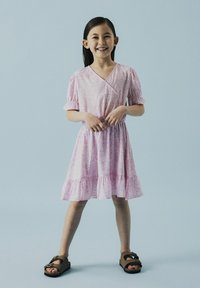 Robe à motifs floraux roses avec un design croisé, manches courtes bouffantes et ourlet volanté. Portée avec des sandales plates marron.