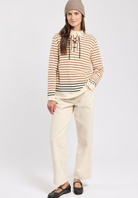 Stribet langærmet top i creme, orange og sort; har en hals snørrebånd og knapdetaljer. Kombineret med cremefarvede brede bukser og sorte sko.