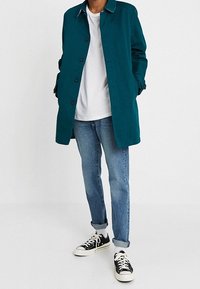 Persona que lleva un abrigo de botones en color turquesa, una camiseta blanca, unos jeans azules arremangados, calcetines blancos y zapatillas negras con cordones blancos.