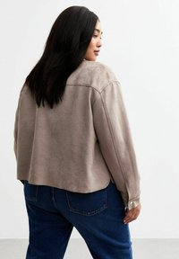 Chaqueta de ante sintético beige con un corte relajado, dobladillo redondeado y puños abotonados, combinada con jeans de mezclilla azul oscuro. Textura suave, con un diseño minimalista.