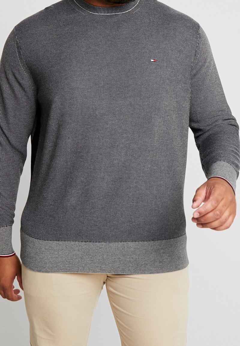 Homme portant un pull à manches longues gris texturé avec des poignets et un ourlet côtelés, associé à un pantalon beige sur un fond neutre.