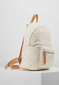 Sac à dos en tissu crème avec un logo à motifs, accents en cuir lisse couleur tan, forme arrondie, poche à zip à l'avant et bretelles réglables.