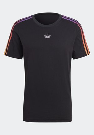 Man draagt zwart Adidas-shirt met kleurrijke strepen, beige cargobroek, zwarte Adidas-sneakers en een zwarte trui over de schouder gehangen.