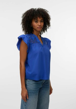 Kobieta o kręconych włosach, ubrana w królewsko niebieską bluzkę z koronkowymi detalami i falbankami na rękawach, zestawioną z jasnoniebieskimi dżinsami.