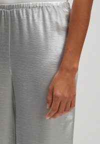 Zijden satijnen broek met een gladde textuur en een elastische tailleband. Een close-up toont een hand die op de stof rust, wat de glans benadrukt.