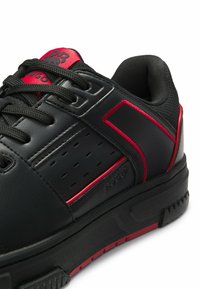 Scarpa da ginnastica nera bassa con dettagli rossi, lacci neri, suola strutturata e collarino imbottito vista di lato.