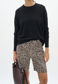 Personne portant un pull noir à manches longues et un short mi-cuisse à imprimé léopard, tenant un sac fourre-tout en cuir marron à côté d'elle.