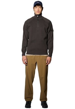 Männliches Model trägt dunkelgrauen Viertelreißverschluss-Pullover, beige Hose mit kleinem Logo, Navy-Cap und schwarze Sneakers, steht vor weißem Hintergrund.