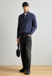Navy zip-neck sweater, black denim jeans, and black shoes. Model holds a small black bag. Simple design with textured fabric and casual fit.