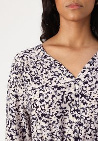 Button-up blouse in light beige with dark blue abstract print. Features a V-neckline, textured fabric, and three buttons down the front.