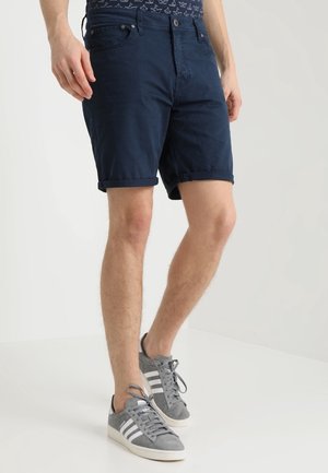 Mand iført mørkeblå opkrogne shorts og grå Adidas-sneakers med hvide striber, der står på en ensfarvet lys baggrund.