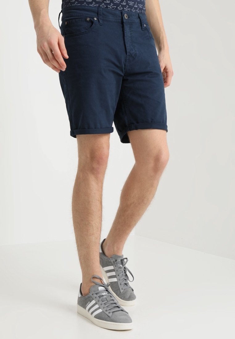 Homme portant un short bleu foncé retroussé et des baskets Adidas grises avec bandes blanches, debout sur un fond clair uni.