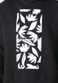 Sudadera negra con capucha que presenta una gran impresión rectangular blanca con patrones florales y el texto "IBASHSTUDIOS" en el centro. Material suave, diseño casual.