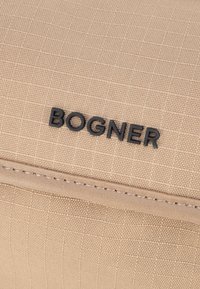 Tessuto beige con texture e lettere in rilievo nere che formano la parola "BOGNER" sopra un bordo in pelle cucito.