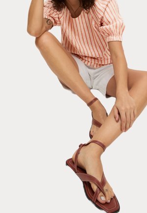 Kvinde siddende med krydsede ben, iført en rød- og hvidstribet top, hvide shorts og brune ankelremssandaler på en ensfarvet baggrund.