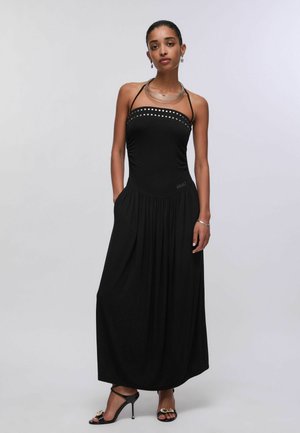 Donna che indossa un abito lungo nero senza maniche con scollo borchiato, mani nelle tasche, collana d'argento, orecchini, braccialetto e sandali neri con tacco alto.