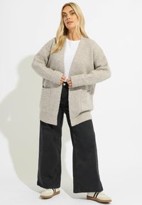 Beige stickad cardigan med lös passform, sänkta axlar och framfickor, kombinerad med svarta vida byxor och krämfärgade sneakers.