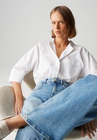 Chemise blanche à boutons avec une poche, assortie à un jean bleu clair à jambes larges. Modèle assis sur une chaise de couleur neutre.