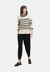 Gestreifter Pullover in Creme und Schwarz mit hochgeschlossenem Kragen und gerippter Textur, kombiniert mit schwarzen, schmal zulaufenden Cargo-Hosen und schwarzen Slippern.