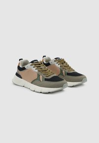 Par de zapatillas bajas en beige, negro y verde oliva con suelas gruesas blancas y cordones marrón claro sobre un fondo gris liso.