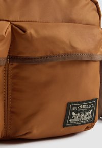 Rudas audinys kuprinė su priekiniais kišenėmis, ant kurios apatinio dešiniojo kampo siuvinėjamas juodai baltas Levi Strauss & Co. logotipo pleistras.