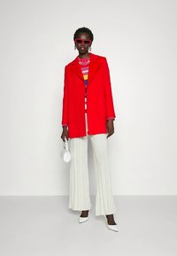 Cappotto rosso in misto lana con rever a incavo sopra maglione a righe multicolore, abbinato a pantaloni bianchi svasati con trama e tacchi a spillo.