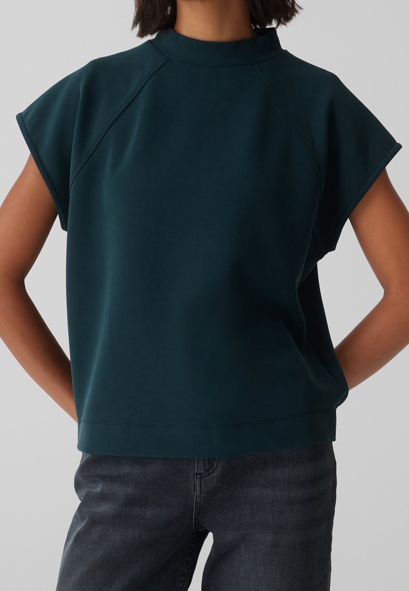 Top a maniche corte color verde acqua scuro con collo alto, realizzato in tessuto liscio; presenta maniche raglan e una vestibilità rilassata.