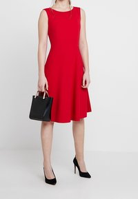 Rode mouwloze jurk met een nauwsluitend lijfje en een uitlopende rok. Gecombineerd met zwarte hoge hakken en een kleine zwarte shopper met goudkleurige accenten.