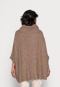 Ruskea neuleponcho, jossa on korkea, taitettu kaulus. Pehmeä rakenne, löysä malli ja leveät hihat, jotka ulottuvat takana puoleen reiteen asti.