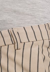 Calças bege com riscas verticais pretas, apresentando um detalhe na cintura e frente pregueada. O tecido tem uma textura suave.