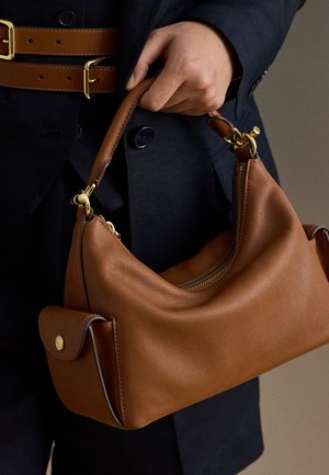 Χέρι κρατά καφέ δερμάτινη τσάντα με χρυσά μεταλλικά στοιχεία, φορεμένη με σκούρο μπλε παλτό που έχει καφέ δερμάτινες ζώνες με χρυσά αγκράφες.