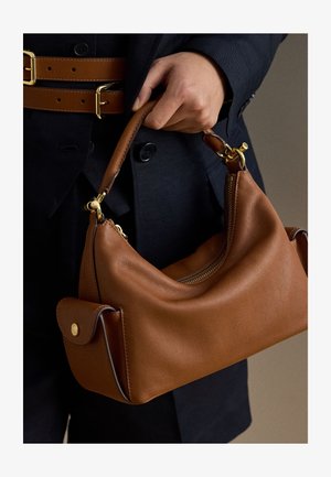 Χέρι κρατά καφέ δερμάτινη τσάντα με χρυσά μεταλλικά στοιχεία, φορεμένη με σκούρο μπλε παλτό που έχει καφέ δερμάτινες ζώνες με χρυσά αγκράφες.