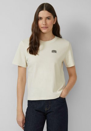 Jeune femme aux longs cheveux bruns portant un t-shirt crème avec le texte "CUTE" et un jean bleu foncé, se tenant devant un fond gris.