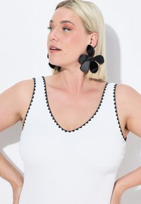 Un débardeur blanc avec un bord festonné noir, affichant un décolleté en V prononcé. De grandes boucles d'oreilles florales noires ajoutent du détail au look.