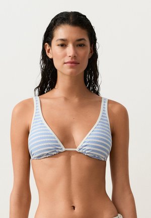 Frau mit nassen Haaren, die einen hellblau-weiß gestreiften Triangel-Bikini-Oberteil trägt, steht vor einem einfarbigen hellen Hintergrund.