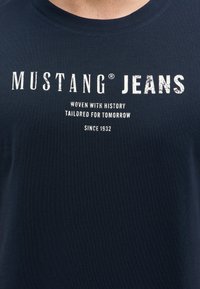 Námořnická modrá pánská tričko s krátkým rukávem vyrobené z bavlny, se strukturovanou tkaninou a bílým potiskem textu: "MUSTANG JEANS," a dalšími detaily níže.