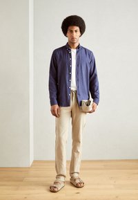 Model wearing a navy button-up shirt, white T-shirt, beige linen pants, and light sandals, holding a light gray clutch bag.